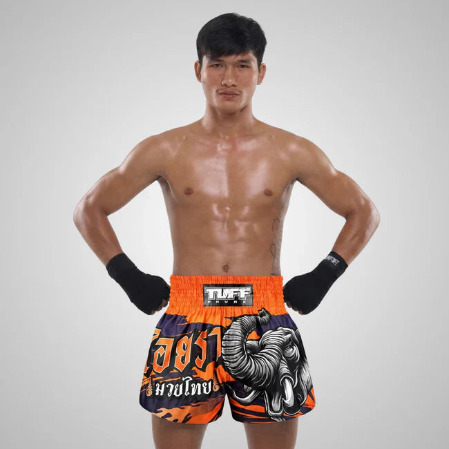 Man wearing orange and black Muay Thai shorts with visible branding on a gray background